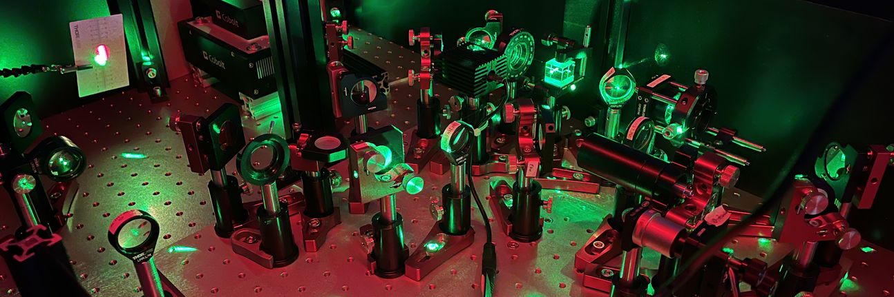 The image shows a laboratory setup with optical components such as lenses, mirrors, and beam splitters. One can see the green laser light reflecting on the components.