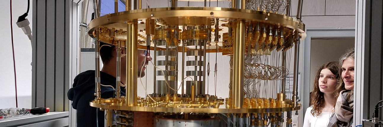 The foreground shows the inner workings of a cryostat for superconducting quantum computing. In the background, a woman and a girl listen to a scientist explaining the cryostat.