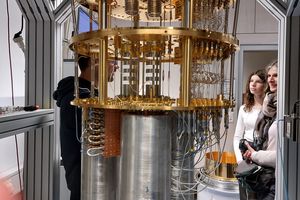 The foreground shows the inner workings of a cryostat for superconducting quantum computing. In the background, a woman and a girl listen to a scientist explaining the cryostat.