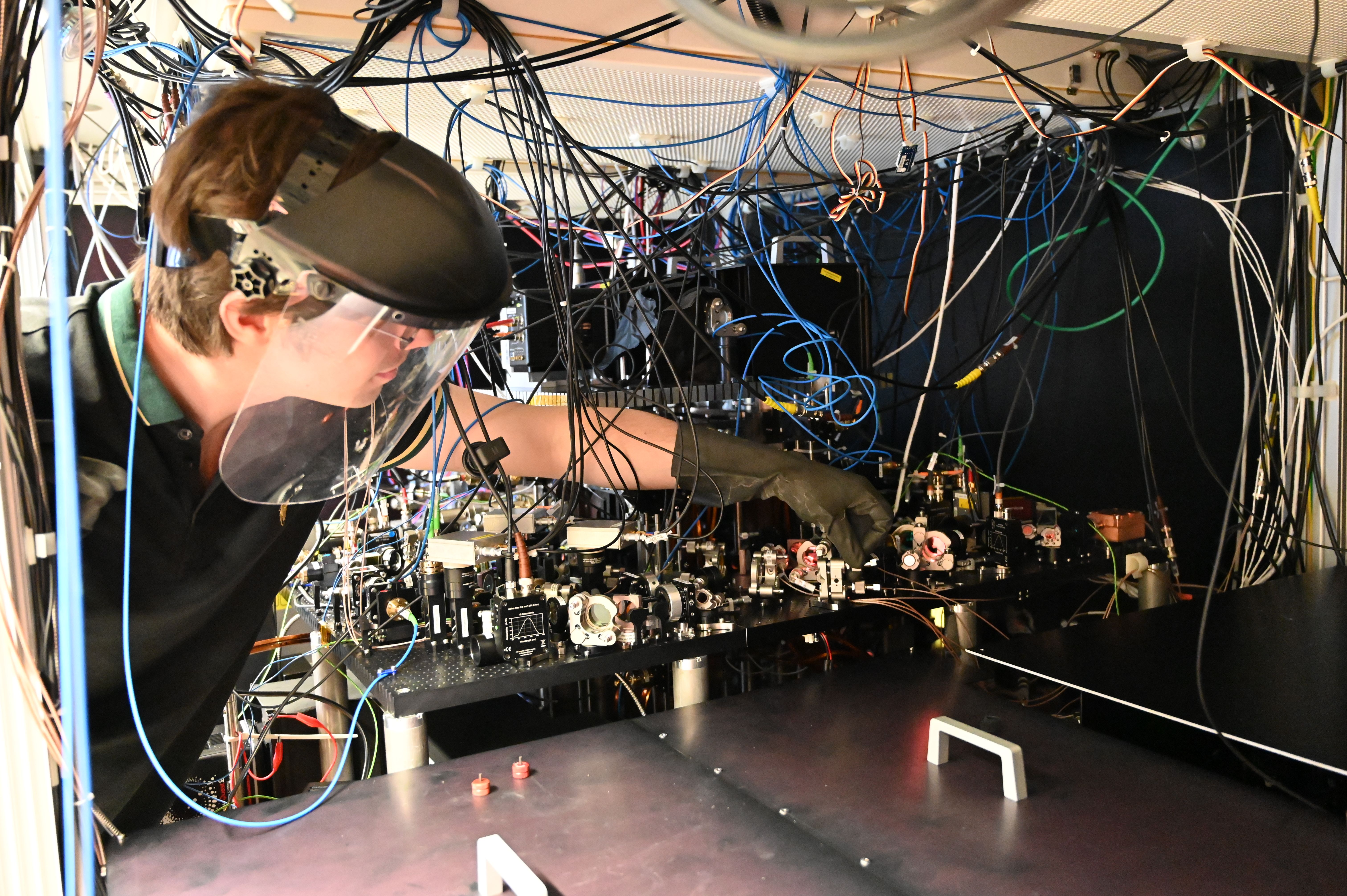 Physicist Dimitris Tsevas is adjusting optical components. Various components, such as mirrors and lenses, can be seen mounted closely together on a plate. Numerous cables run from the individual components up to the ceiling. Dimitris is wearing a face shield and protective gloves.