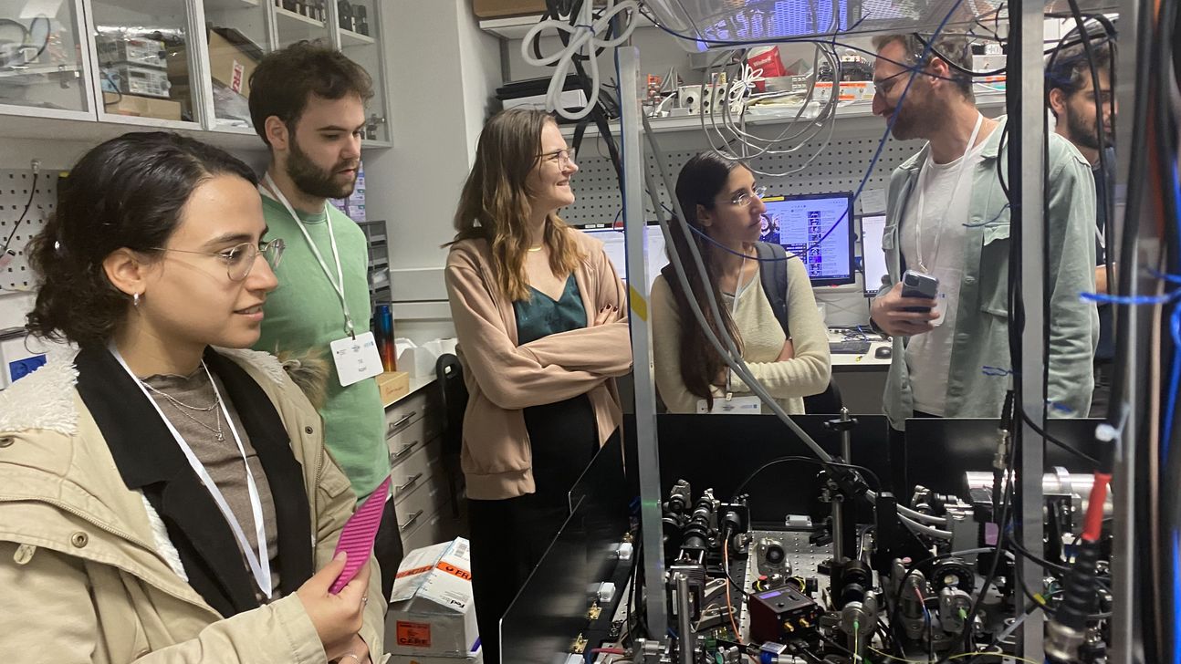 Students visiting a quantum lab. They stand at a table with measuring devices.