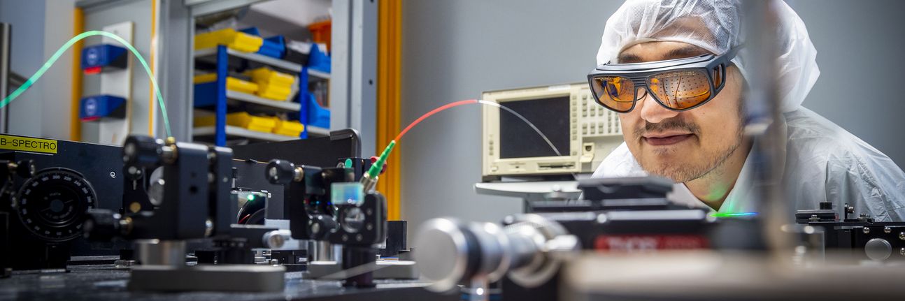 A scientist with laser googles and a hairnet examines an optical experimental setup.