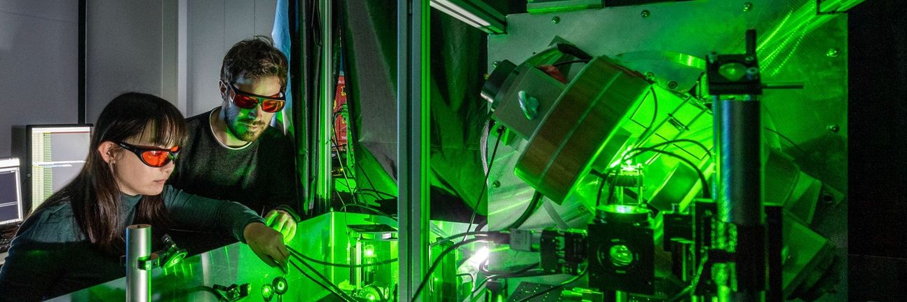 A woman and a man are standing at an experimental table. They are wearing protective goggles with red glass. Green laser light is radiating in the experiment.