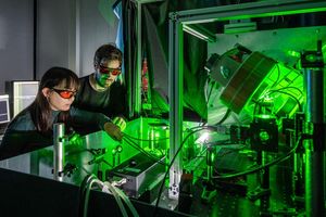 A woman and a man are standing at an experimental table. They are wearing protective goggles with red glass. Green laser light is radiating in the experiment.