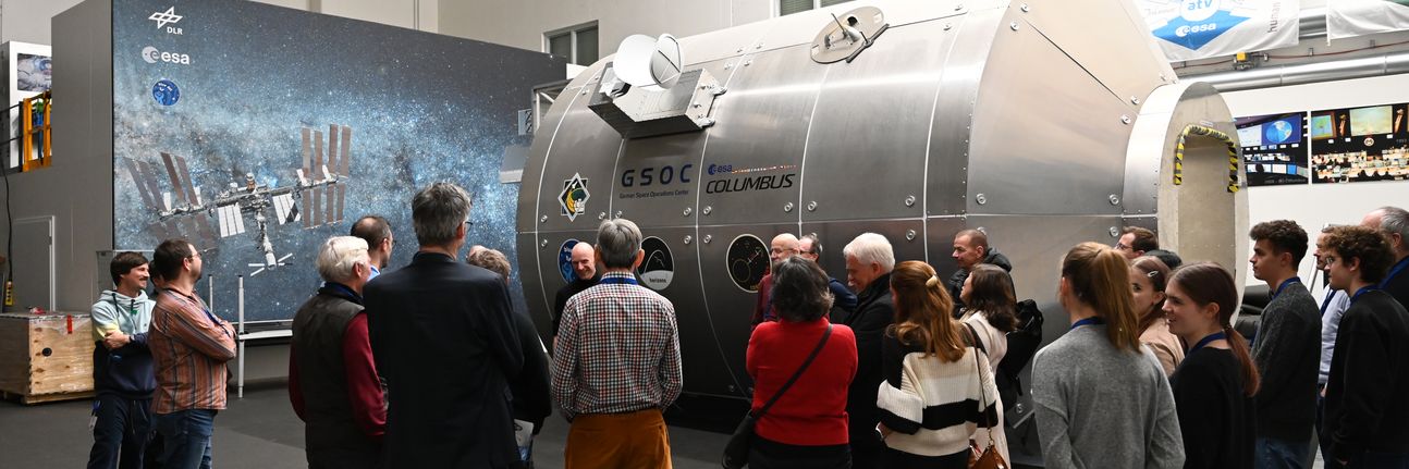 A group of people listen to a scientist from the German Aerospace Center. In the background, one can see a model of the Columbus module.