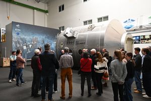 A group of people listen to a scientist from the German Aerospace Center. In the background, one can see a model of the Columbus module.
