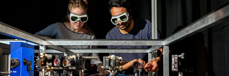 A young female scientist and her colleague look at an optical experiment wearing laser googles.