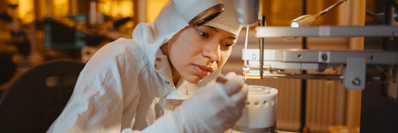 A female researcher in a cleanroom suit uses tweezers to place a material sample under a microscope.
