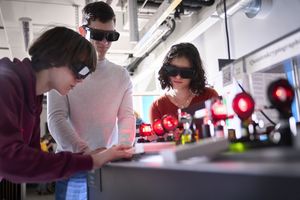 Two boys and a girl are wearing laser goggles and conducting an experiment with laser light. You can see the red laser light passing through several polarization filters.