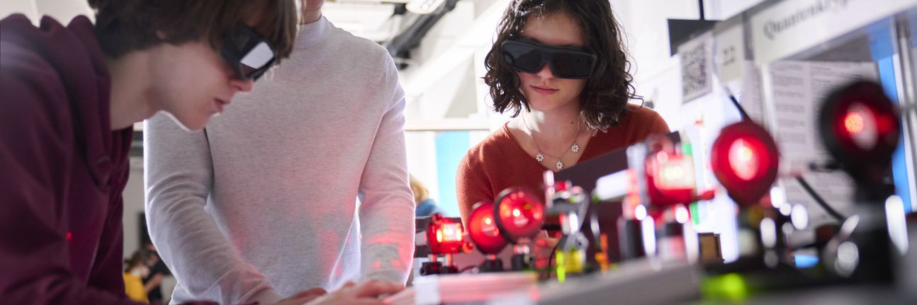 Two boys and a girl are wearing laser goggles and conducting an experiment with laser light. You can see the red laser light passing through several polarization filters.