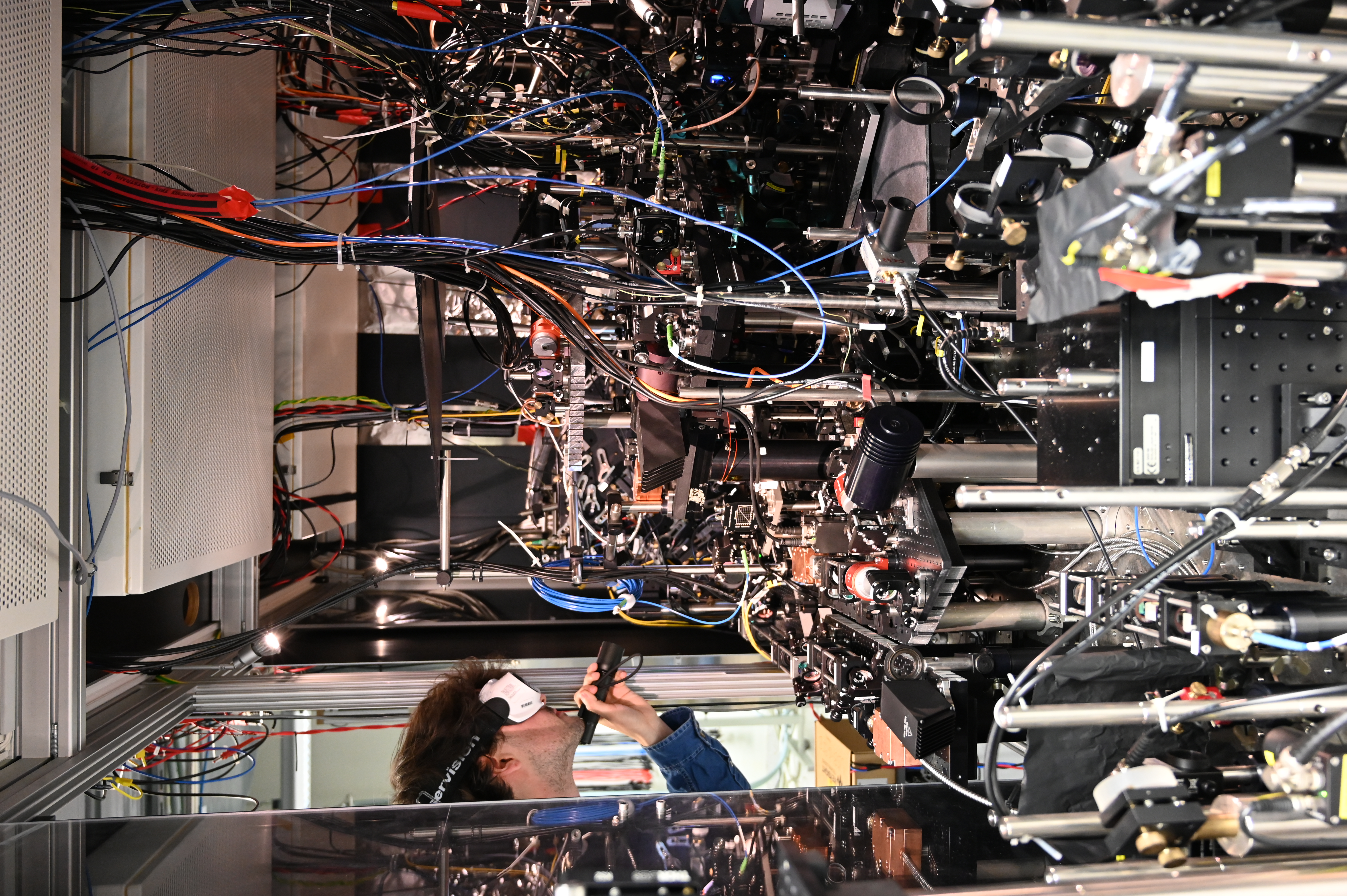 The physicist wears laser goggles and, equipped with a flashlight, observes the experimental set-up. Numerous optics and cables are piled up on several levels.