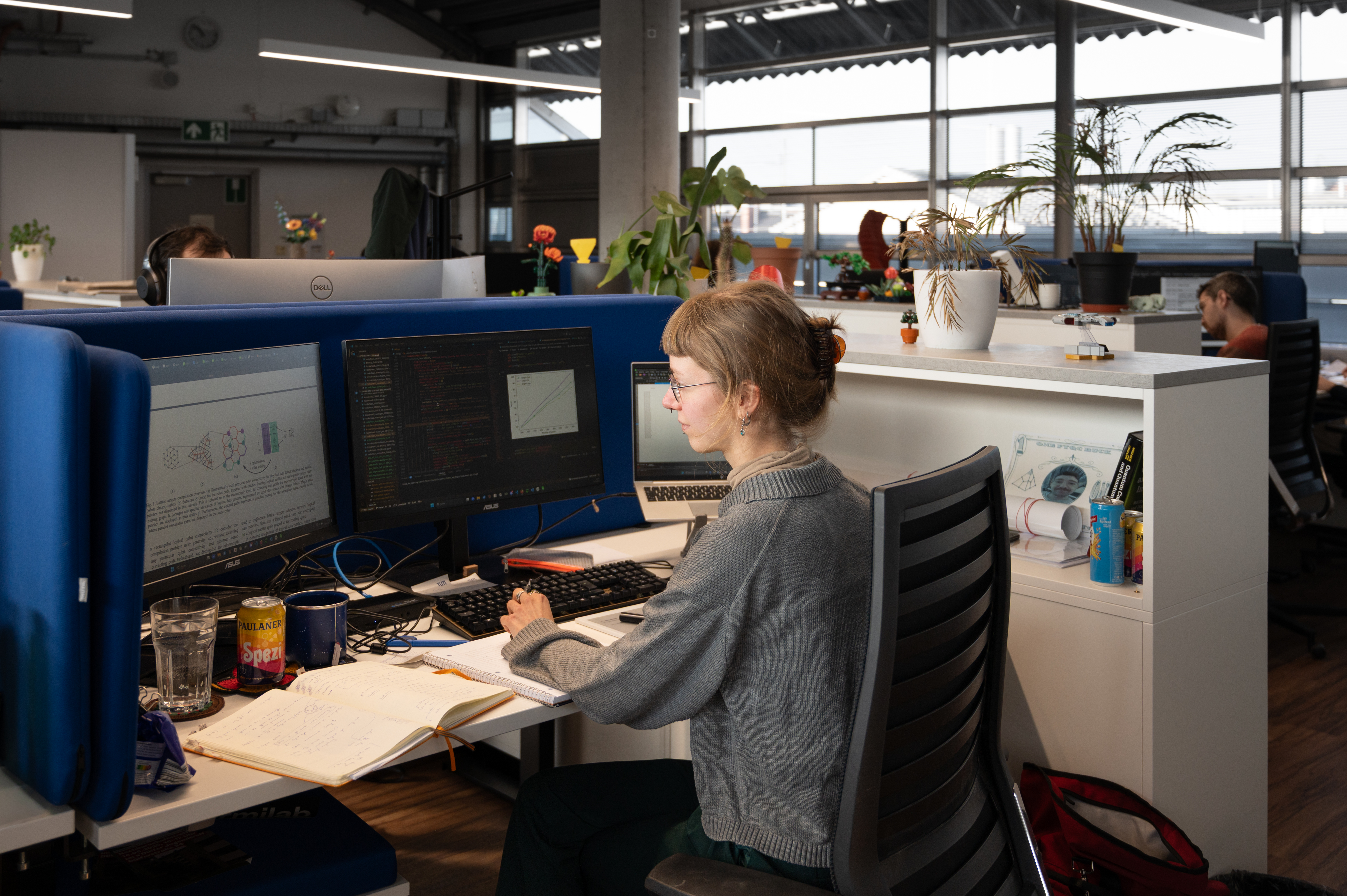 Laura Herzog is sitting at her desk with her can of “Spezi” soda. The open-plan office can be seen in the background. Her colleagues are sitting at the other desks. The room is decorated with plants and colourful Lego flowers.