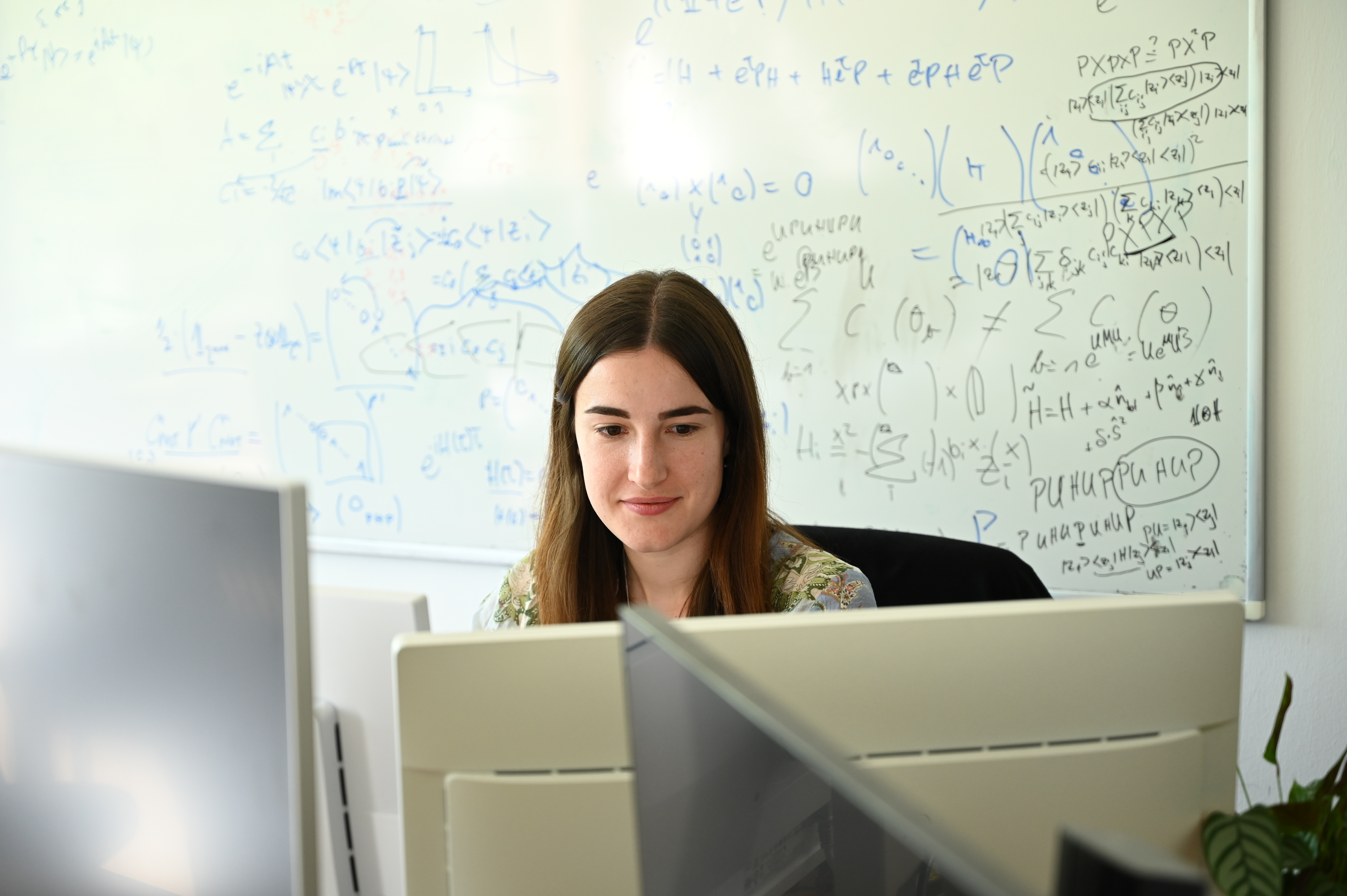 A scientist sits at a desk, looking intently at a computer screen. In the background, there is a whiteboard covered in formulas.