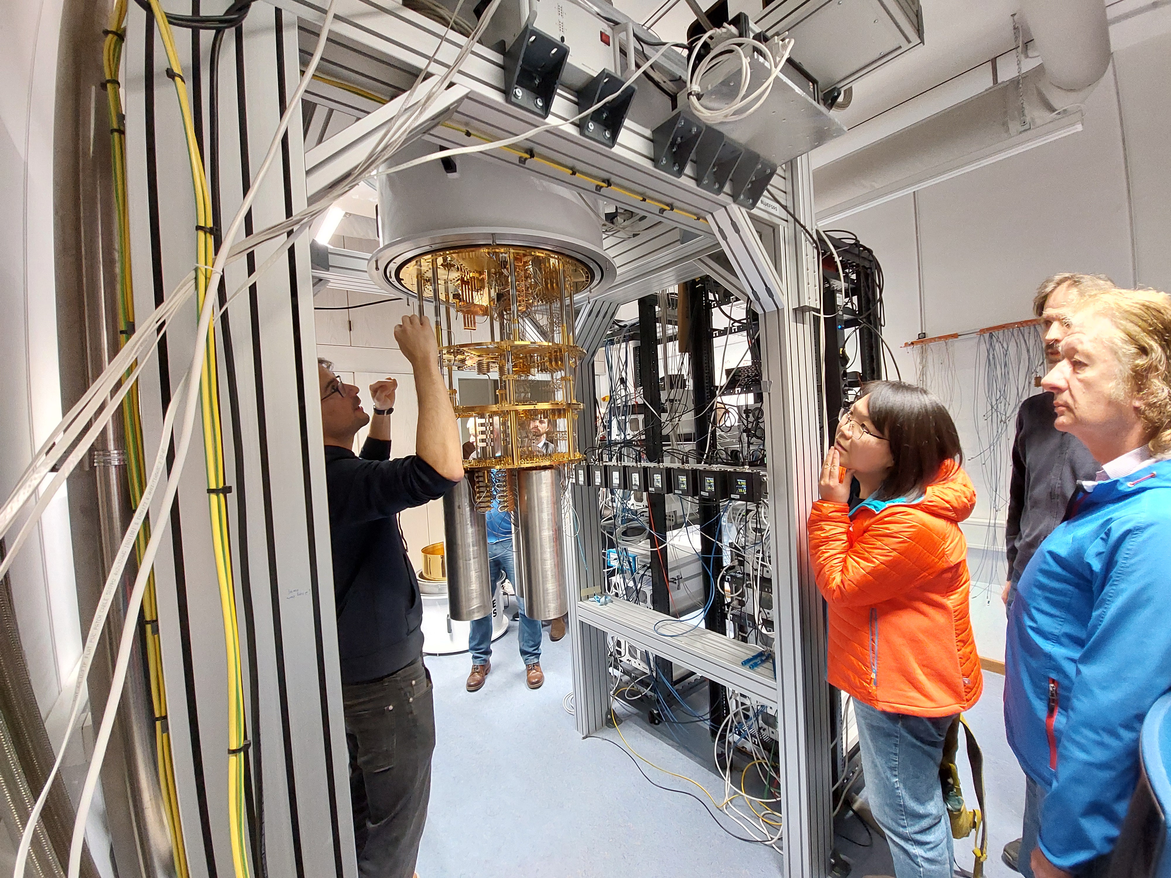 A scientist and several visitors look with interest at the inner workings of a cryostat for superconducting quantum computing. They discuss the various components of the cryostat.