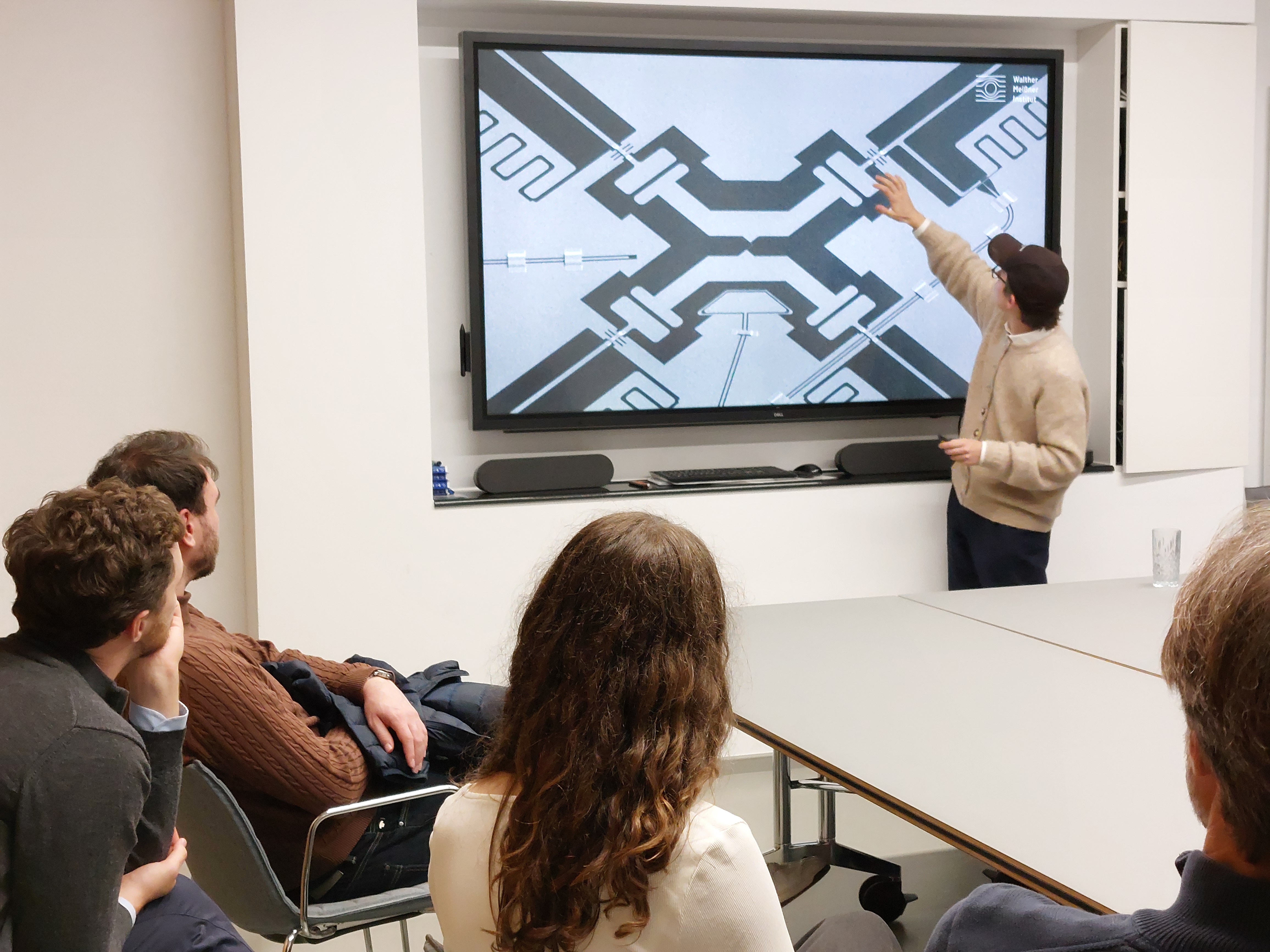 A young scientist gives a presentation and points to an element of an image showing a superconducting qubit. People sit in the foreground listening to him.